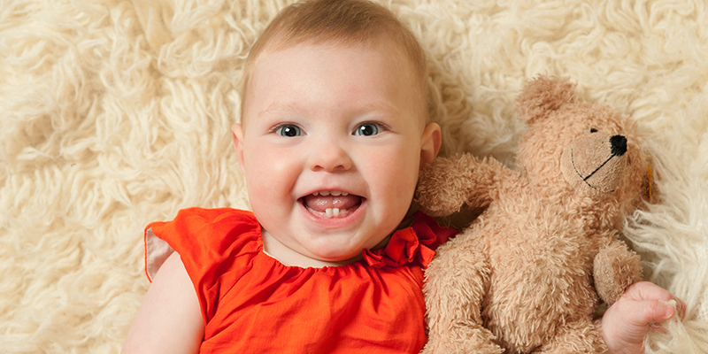 nine month old girl in orange dress cuddling teddy and smiling at the camera