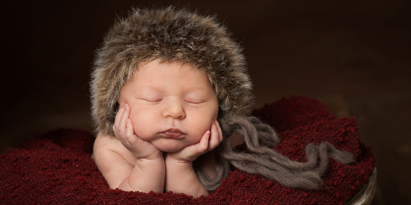 newborn girl in furry bonnet on red blanket in the froggy pose